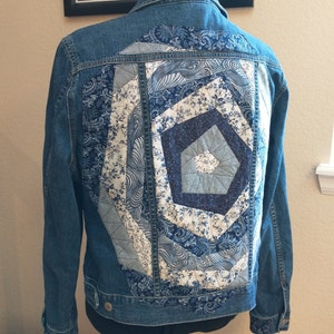 May include: A blue denim jacket with a patchwork design on the back. The patchwork is made of different blue and white fabrics with floral and geometric patterns.