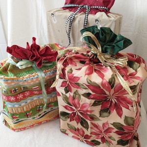 May include: Three fabric gift bags with decorative tops. One bag has a red and white floral pattern, another has a green and red pattern, and the third has a beige background with snowmen. Each bag is tied with ribbons.