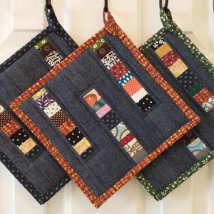May include: Three quilted pot holders in shades of blue, orange, and green hang from hooks. Each square pot holder features a patchwork design with various fabric patterns and colors, including floral and polka dots.