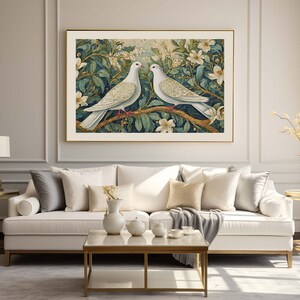 May include: A white couch with gray and white throw pillows sits in front of a wall with a gold frame. The frame contains a print of two white doves perched on a branch with green leaves and white flowers.