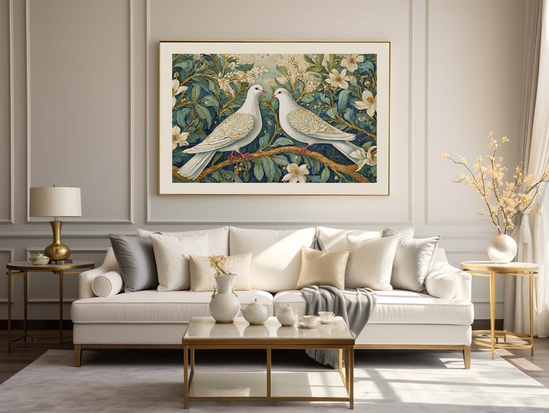 May include: A white couch with gray and white throw pillows sits in front of a wall with a gold frame. The frame contains a print of two white doves perched on a branch with green leaves and white flowers.