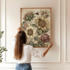 Puede incluir: Un estampado bot&aacute;nico enmarcado con girasoles, alcachofas y otras flores en tonos apagados. La obra de arte incluye una abeja y est&aacute; sostenida por una persona. El marco es de madera clara.