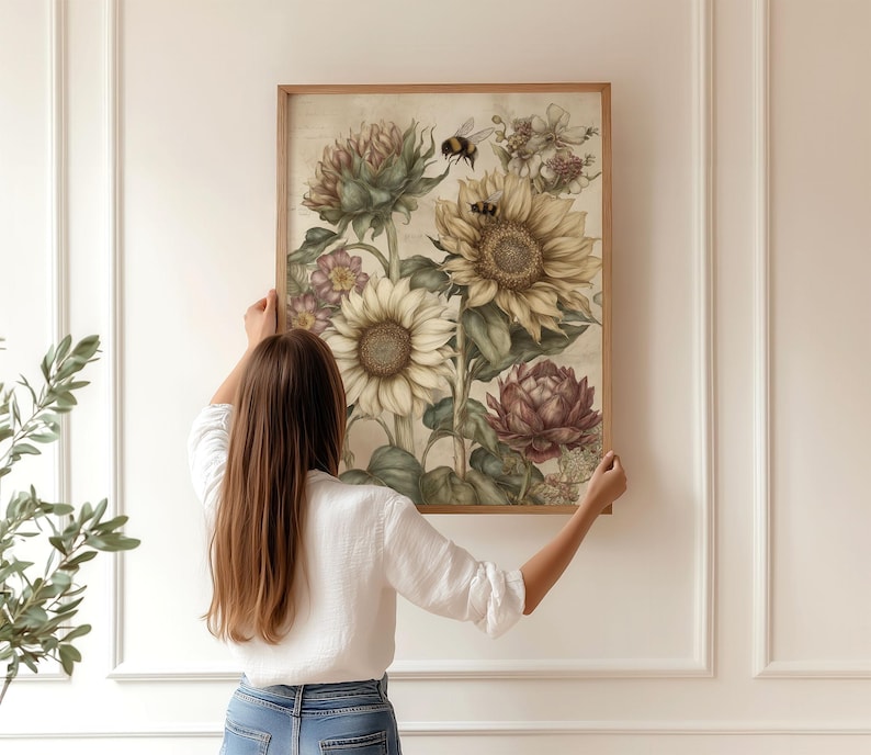 Puede incluir: Un estampado bot&aacute;nico enmarcado con girasoles, alcachofas y otras flores en tonos apagados. La obra de arte incluye una abeja y est&aacute; sostenida por una persona. El marco es de madera clara.