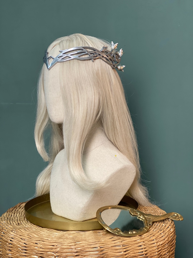 Puede incluir: Corona plateada con dise&ntilde;o de hojas, puesta en una cabeza de maniqu&iacute; blanca con cabello largo y rubio. El maniqu&iacute; est&aacute; sentado en una superficie de mimbre con una bandeja dorada y un peque&ntilde;o espejo de mano.