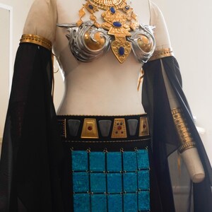 May include: A costume featuring a gold and silver chest piece with blue and red accents, a matching choker, and arm cuffs. The outfit includes a black skirt with a central panel of shimmering blue square tiles and a belt with gold and black geometric details.