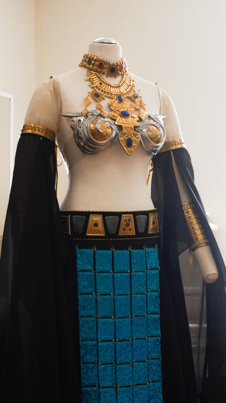 May include: A costume featuring a gold and silver chest piece with blue and red accents, a matching choker, and arm cuffs. The outfit includes a black skirt with a central panel of shimmering blue square tiles and a belt with gold and black geometric details.
