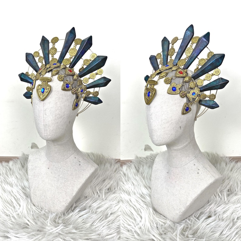 May include: A decorative headpiece featuring a gold-toned metal frame adorned with dark blue, pointed, crystalline-shaped spikes. The structure includes small gold circular medallions, heart-shaped charms with blue and red gems, and a wire base.