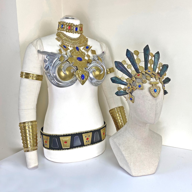 May include: A costume set featuring a gold and silver chest piece with blue and red gems, a matching choker, arm cuffs, and a black belt with gold accents. Beside it, a headpiece with dark blue crystal-like spikes and gold filigree details.