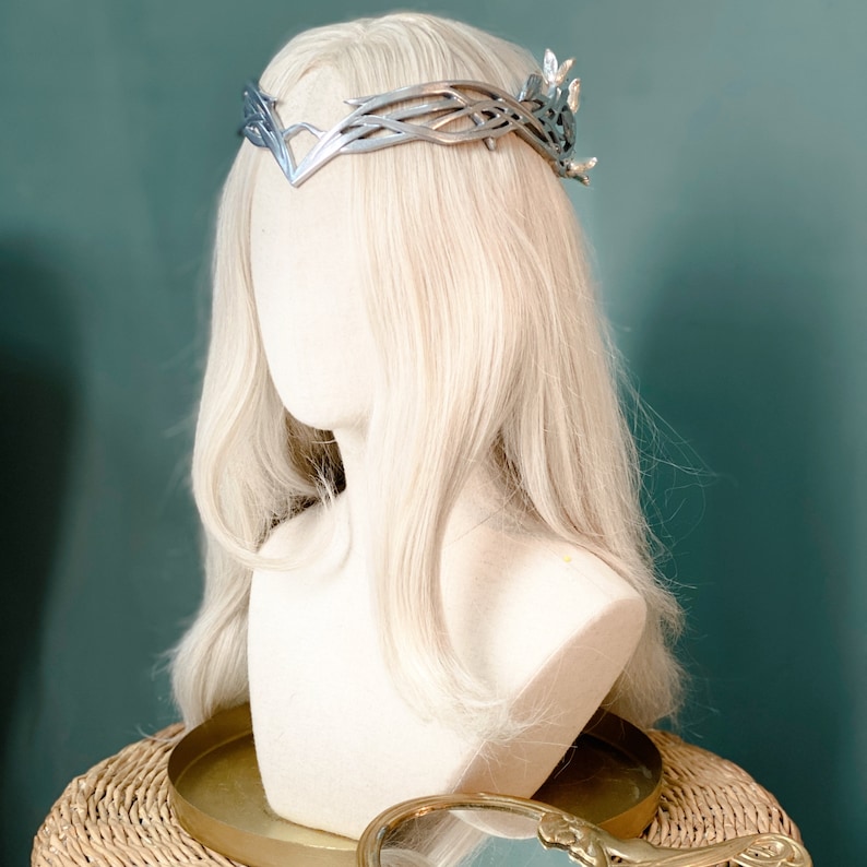 Puede incluir: Una cabeza de maniqu&iacute; blanca con cabello largo y rubio platino que lleva una corona plateada con un dise&ntilde;o de hojas.