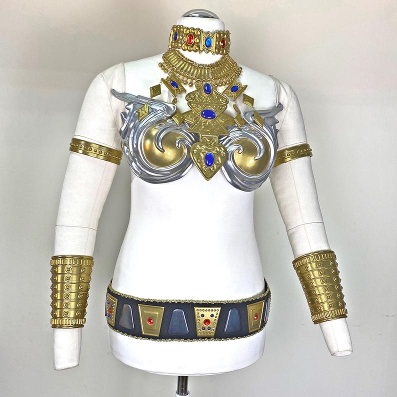 May include: A costume set featuring a gold and silver chest piece with blue accents, a matching gold choker necklace, two gold upper arm bands, two gold forearm cuffs, and a black belt with gold geometric plates and red and blue stone inlays.