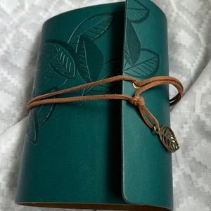 May include: A dark green leather journal with a leaf design embossed on the cover. The journal has a brown leather strap with a metal leaf charm.