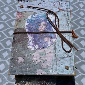 Könnte beinhalten: Ein handgemachtes Journal mit einem blauen und weißen gemusterten Einband. Der Einband zeigt einen Aufkleber einer Frau mit langen violetten Haaren. Das Journal ist mit einer braunen Lederschnur zugebunden.