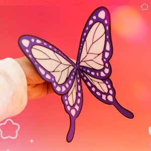 May include: A flat, two-dimensional butterfly cutout featuring a vibrant purple border with white circular accents and white inner wing sections defined by black outlines. The butterfly has long, slender lower wing tails and a symmetrical shape.