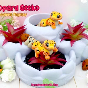 Può includere: Una fioriera bianca a più livelli per piante grasse con una statuina di geco leopardo dei cartoni animati. La fioriera ha finte piante grasse rosse e verdi. Il testo "Leopard Gecko & Succulent Planter" è in alto. Il testo "Downloadable STL Files" è in basso.