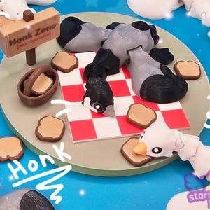 Puede incluir: Una figurita de ganso gris y negro con una manta de picnic a cuadros rojos y blancos, rodeada de pequeñas rebanadas de pan marrón y un cartel que dice "Honk Zone (No Humans!)".