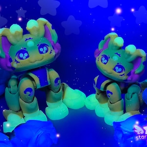 May include: Two articulated, glowing figurines with yellow, green, and purple accents. The figures have large eyes and are posed against a dark blue background with star shapes. The figures are sitting on a blue surface. The Star Mouse 3D logo is visible.