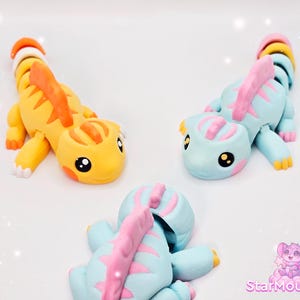 Può includere: Tre figurine di lucertola colorate in argilla polimerica fatte a mano. Una è gialla con strisce arancioni, le altre due sono azzurre con dettagli rosa. Ognuna ha grandi occhi neri e una coda segmentata. Il logo StarMouse3D è visibile in basso a destra.