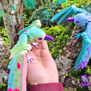 Puede incluir: Dos coloridas figuras de pájaros, en tonos azules, verdes y rosas, posadas en una rama de árbol. Los pájaros tienen picos y patas moradas, con un diseño caprichoso. El fondo presenta un exuberante follaje verde y un toque de brillo.