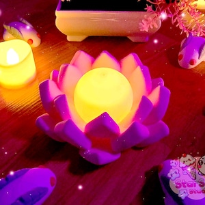 May include: A purple lotus flower candle holder with a yellow LED candle inside. The candle holder is surrounded by small, white, ceramic fish figurines. The background is a dark wood surface.
