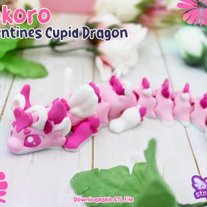 Może przedstawiać: Różowa i biała figurka smoka Kupidyna z napisem "Kokoro Valentines Cupid Dragon". Smok ma akcenty w kształcie serca i białe skrzydła. Figurka stoi na białej drewnianej powierzchni z kwiatami.