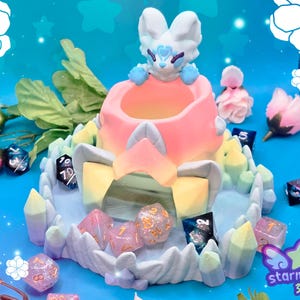 May include: A fantasy-themed dice holder featuring a white and blue creature atop a pink and yellow structure. The holder is surrounded by colorful dice and crystal-like accents. The background is a vibrant blue with floral decorations.