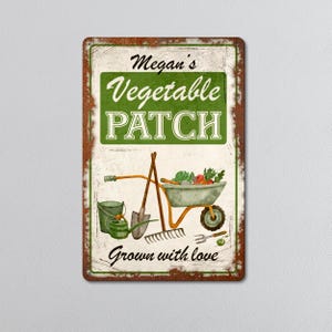 May include: A rectangular metal sign with a distressed, rusty brown border. The sign features the text "Megan's Vegetable Patch" in white and green, along with illustrations of gardening tools and a wheelbarrow filled with vegetables. The words "Grown with love" are also included.
