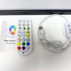 Op de afbeelding: Een witte afstandsbediening met een verscheidenheid aan knoppen, waaronder snelheid, modus en kleurkeuzes. De afstandsbediening is gelabeld "2.4G music controller". Er is ook een witte USB-ontvanger en een witte handleiding.
