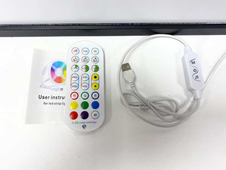 Op de afbeelding: Een witte afstandsbediening met een verscheidenheid aan knoppen, waaronder snelheid, modus en kleurkeuzes. De afstandsbediening is gelabeld "2.4G music controller". Er is ook een witte USB-ontvanger en een witte handleiding.