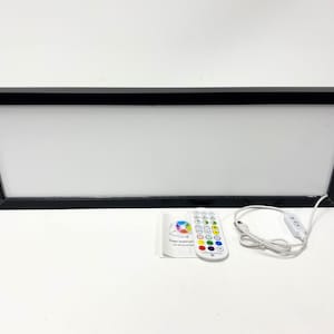 Op de afbeelding: Een zwarte, ingelijste lichtbox met een wit oppervlak en een afstandsbediening. De afstandsbediening heeft verschillende gekleurde knoppen en een aan / uit-knop. De lichtbox is via een wit snoer verbonden met de afstandsbediening.