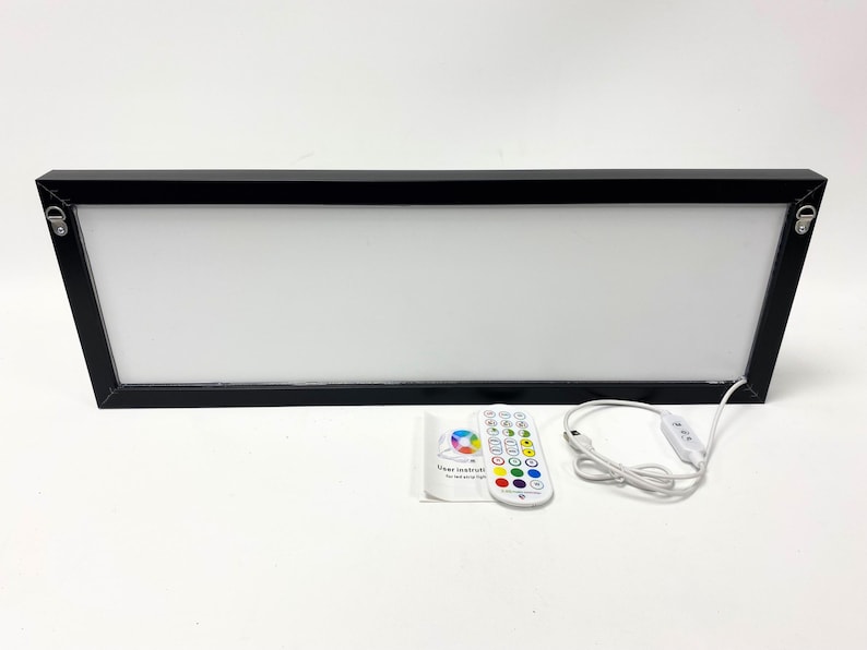 Op de afbeelding: Een zwarte, ingelijste lichtbox met een wit oppervlak en een afstandsbediening. De afstandsbediening heeft verschillende gekleurde knoppen en een aan / uit-knop. De lichtbox is via een wit snoer verbonden met de afstandsbediening.