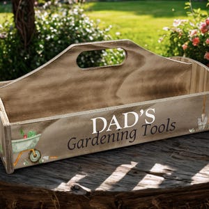 May include: Wooden gardening tool box with a handle, featuring the text "DAD'S Gardening Tools" in white. The box has illustrations of gardening tools and a wheelbarrow. The box is sitting on a wooden surface.