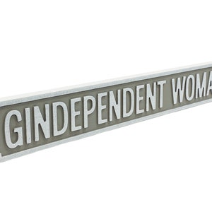 May include: A white and gray sign with the text "GINDEPENDENT WOMAN" in white lettering.