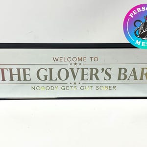 May include: A black framed sign with a white background and gold lettering that reads "Welcome to The Glover's Bar Nobody Gets Out Sober".