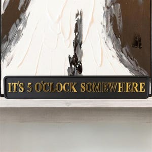 May include: A black sign with gold lettering reading "IT'S 5 O'CLOCK SOMEWHERE". The sign is set against a backdrop of a black and white abstract painting.