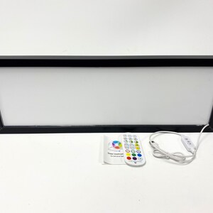 May include: A black framed light box with a white surface and a remote control. The remote control has a variety of color buttons and a power button. The light box is connected to the remote control by a white cord.