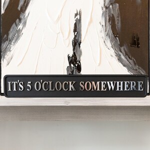 May include: A black rectangular sign with the text "IT'S 5 O'CLOCK SOMEWHERE" in silver lettering. The sign is placed on a white shelf, with a black and white abstract painting in the background.