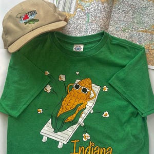May include: A green t-shirt with a graphic of a corn cob wearing sunglasses, relaxing on a white lounge chair. The word "Indiana" is printed below the graphic. A tan baseball cap with "I love fishing" embroidery and a map are also in the image.