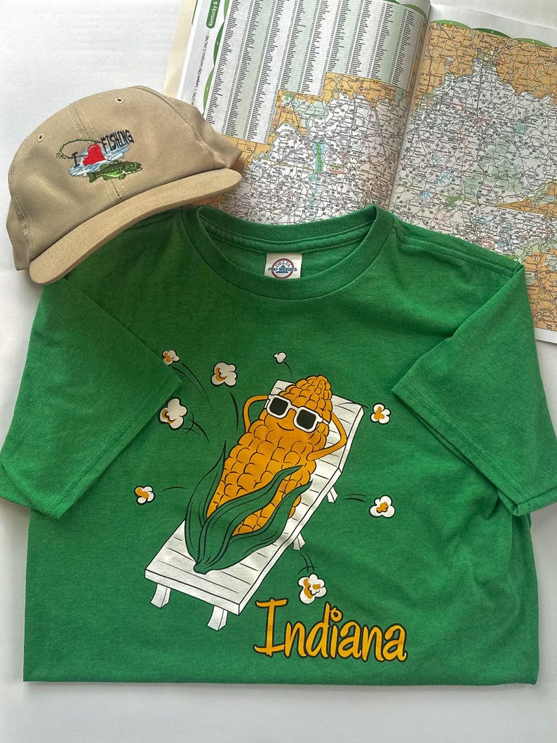 May include: A green t-shirt with a graphic of a corn cob wearing sunglasses, relaxing on a white lounge chair. The word "Indiana" is printed below the graphic. A tan baseball cap with "I love fishing" embroidery and a map are also in the image.