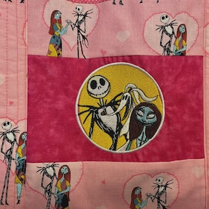 May include: A pink quilted fabric featuring a repeating pattern of Jack Skellington and Sally inside hearts. A central dark pink rectangular panel displays a circular embroidered patch of Jack Skellington, Sally, and Zero against a yellow moon.