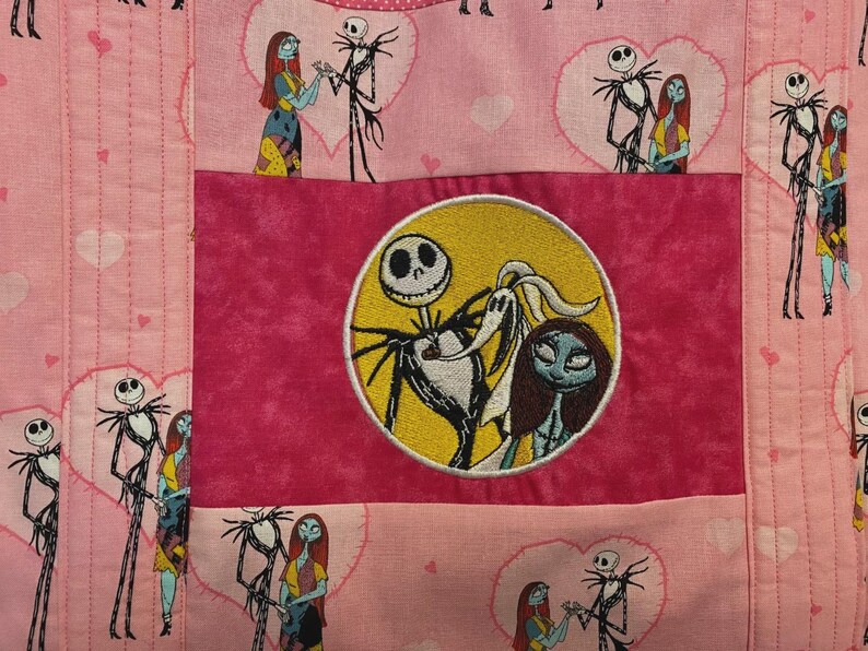 May include: A pink quilted fabric featuring a repeating pattern of Jack Skellington and Sally inside hearts. A central dark pink rectangular panel displays a circular embroidered patch of Jack Skellington, Sally, and Zero against a yellow moon.