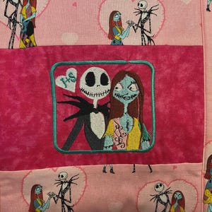 May include: A quilted fabric featuring a repeating pattern of Jack Skellington and Sally from The Nightmare Before Christmas inside pink hearts. A central dark pink rectangular patch displays an embroidered image of the pair with a heart labelled J+S.