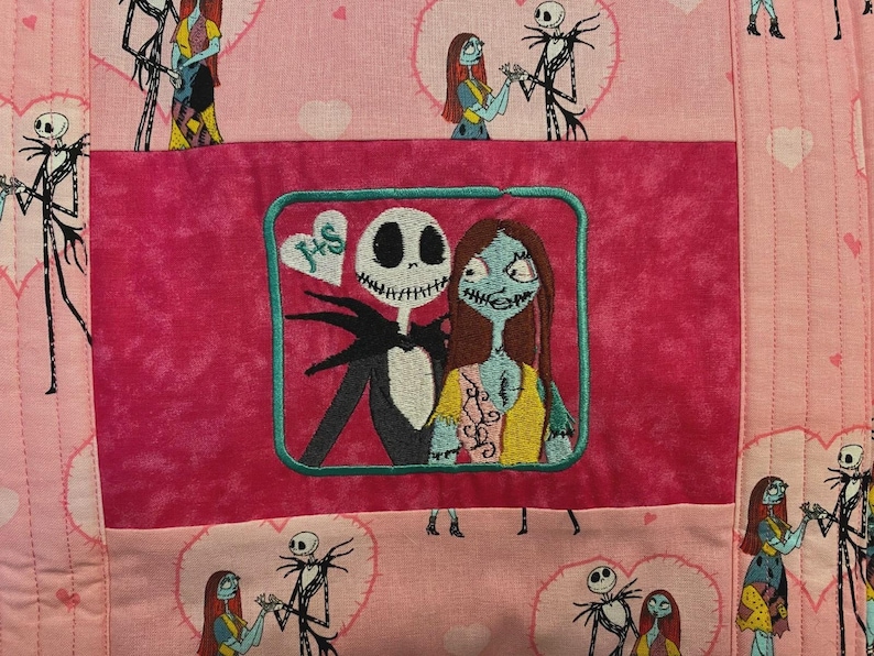 May include: A quilted fabric featuring a repeating pattern of Jack Skellington and Sally from The Nightmare Before Christmas inside pink hearts. A central dark pink rectangular patch displays an embroidered image of the pair with a heart labelled J+S.