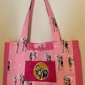May include: A pink tote bag featuring a repeating pattern of Jack Skellington and Sally from The Nightmare Before Christmas inside heart shapes. The bag has a central rectangular patch with a circular image of the characters and side panels.