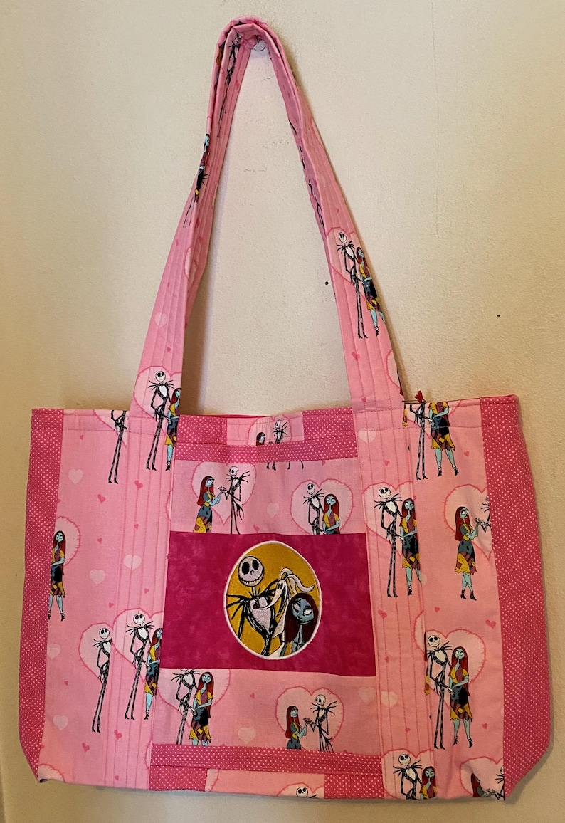 May include: A pink tote bag featuring a repeating pattern of Jack Skellington and Sally from The Nightmare Before Christmas inside heart shapes. The bag has a central rectangular patch with a circular image of the characters and side panels.