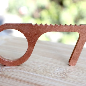 May include: A copper-colored door opener tool with a loop on one end and a serrated edge on the other. The tool is designed to help people avoid touching surfaces in public places.
