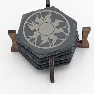 May include: A set of hexagonal slate coasters with a sun and bird design, resting in a dark wood holder. The coasters are dark gray with a lighter gray etched design. The holder has a simple, geometric design.