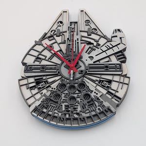 Può includere: Un orologio da parete in legno a forma di astronave Millennium Falcon. L'orologio presenta un design dettagliato e stratificato con elementi grigi e neri, lancette rosse e un accento blu. È montato su una parete bianca.