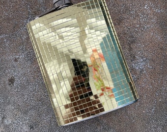 May include: A rectangular gold flask with a mirrored tile design. The flask has a silver cap and is set against a textured, dark gray surface. The mirrored surface reflects the surrounding environment.
