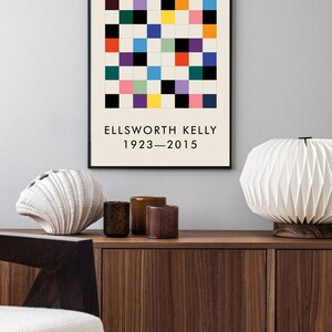 May include: A black framed print with a colorful geometric pattern and the text "ELLSWORTH KELLY 1923-2015". The print is hanging on a light gray wall above a wooden cabinet with two drawers.