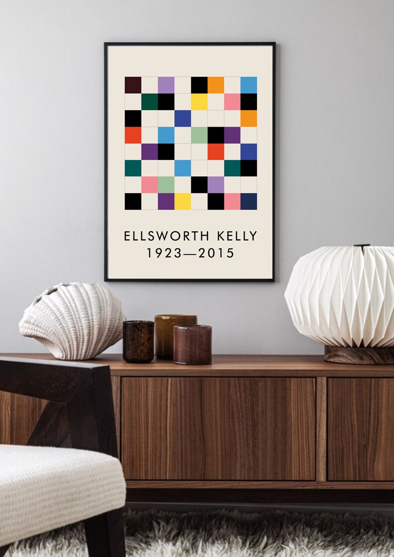 May include: A black framed print with a colorful geometric pattern and the text "ELLSWORTH KELLY 1923-2015". The print is hanging on a light gray wall above a wooden cabinet with two drawers.